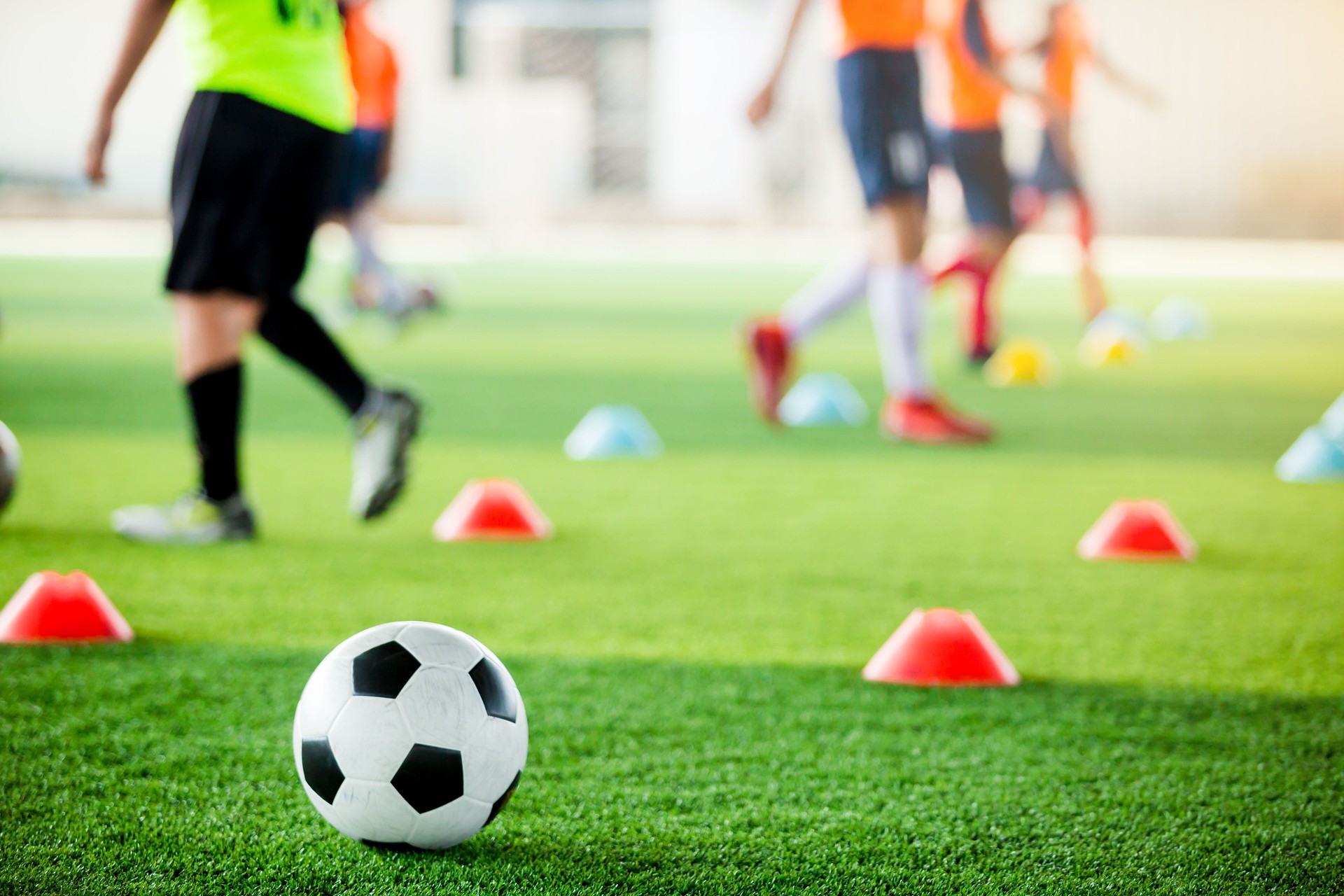 football on green artificial turf with blurry soccer team training, blurry kid soccer player jogging between marker cones and control ball