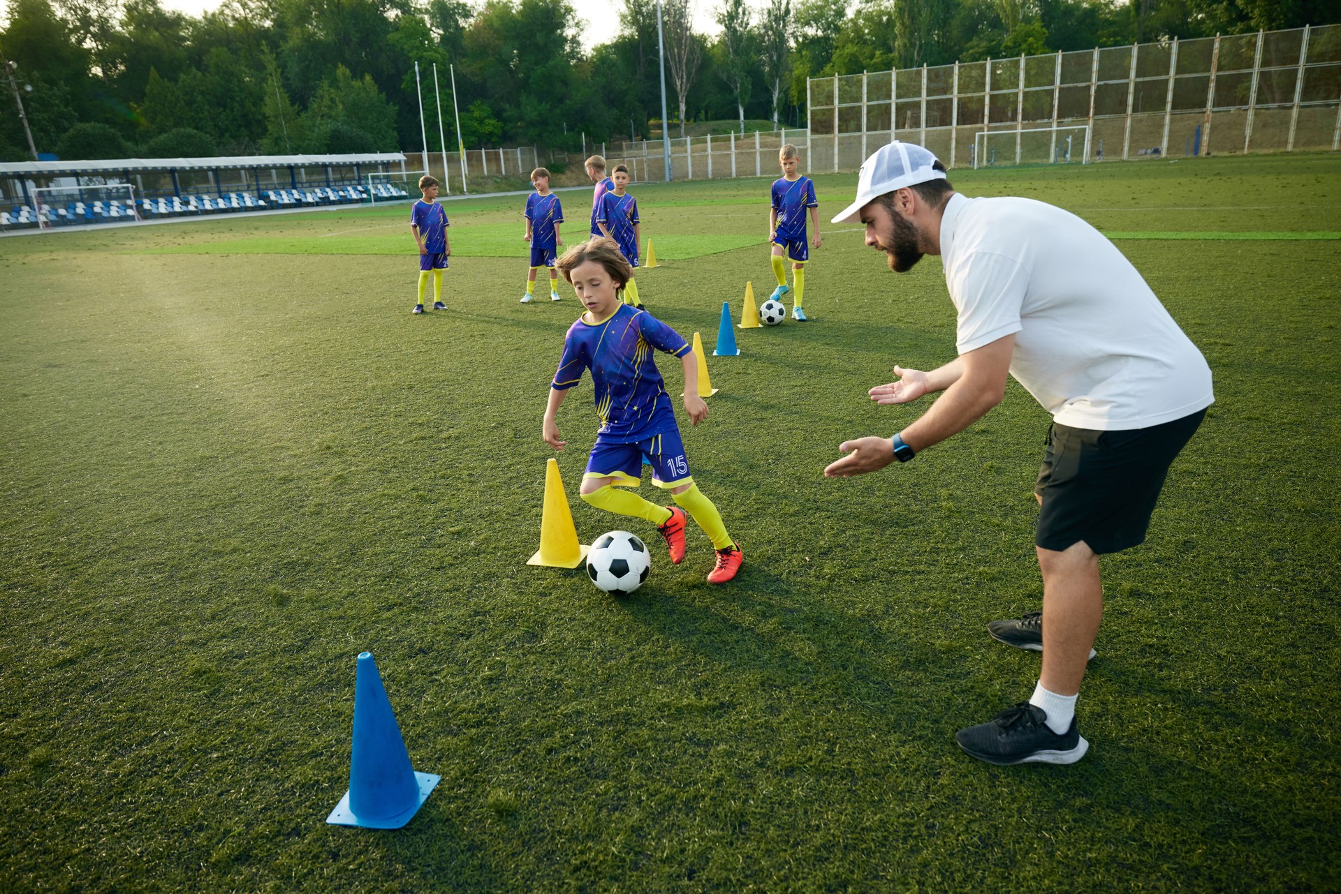 Boy practices dribbling through cones with focused encouragement from the coach. Outdoor stadium and evening game.
