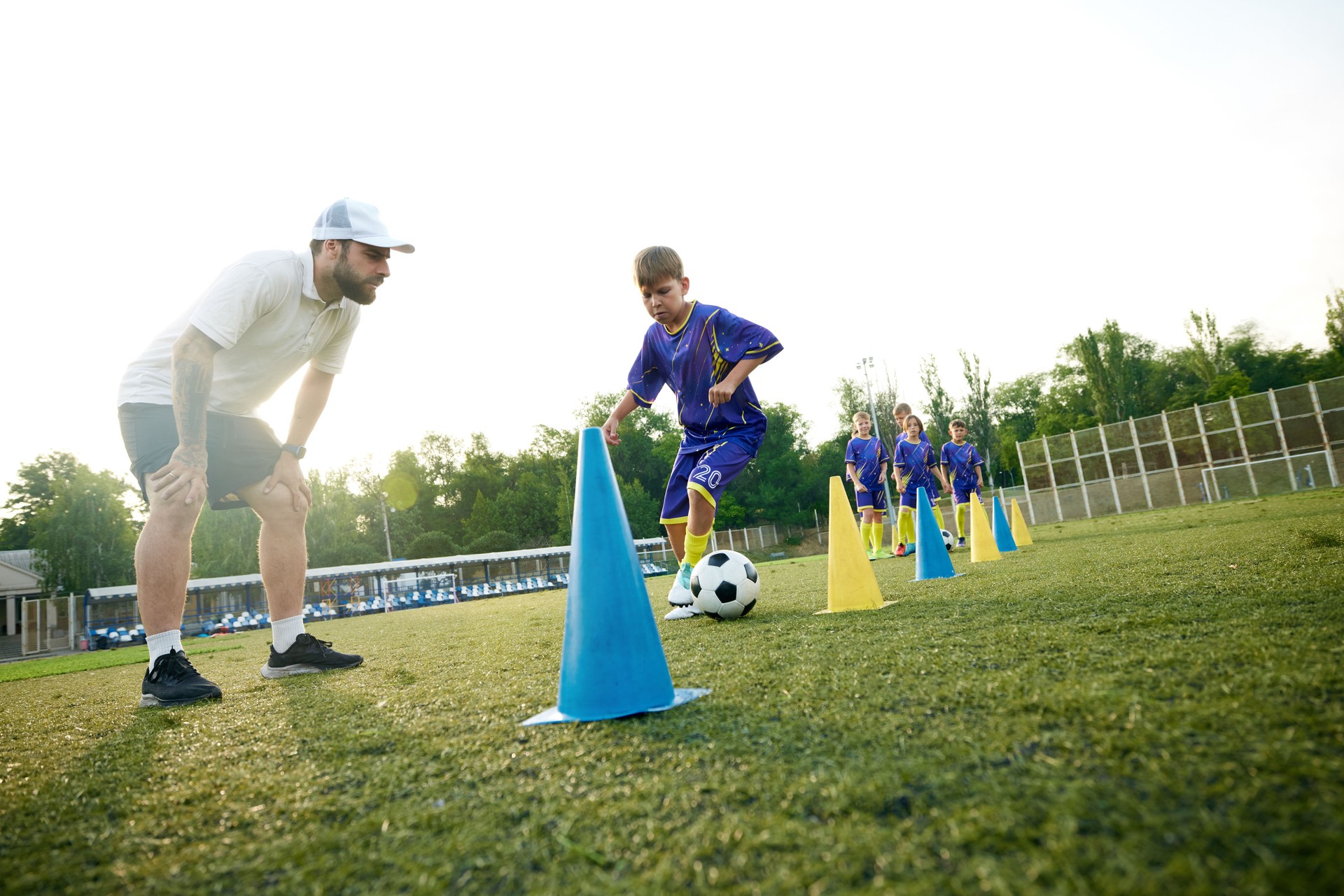 Young boy, child in motion, dribbling soccer ball around cones and receiving guidance from coach. Outdoor stadium game